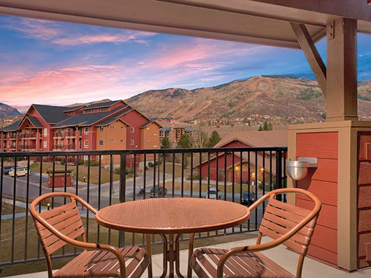 Private outdoor balcony with a table and two chairs overlooking the Steamboat Springs resort and mountains under a colorful sunset sky.