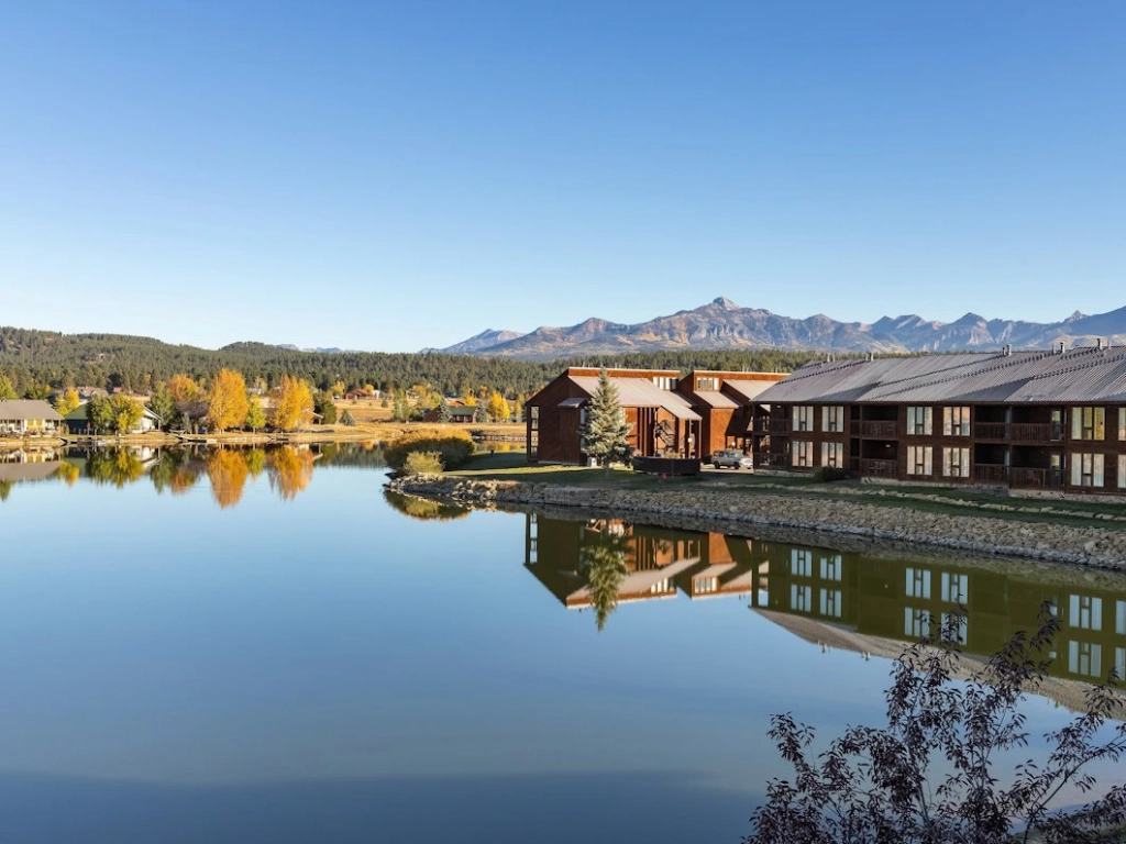 Serene lakeside view of Pagosa Springs resort buildings reflecting on the calm water with mountain peaks in the distance, capturing the essence of summer getaways in Colorado.