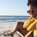 Smiling woman wearing orange sunglasses relaxing in a wooden beach chair on a sunny beach by the ocean.