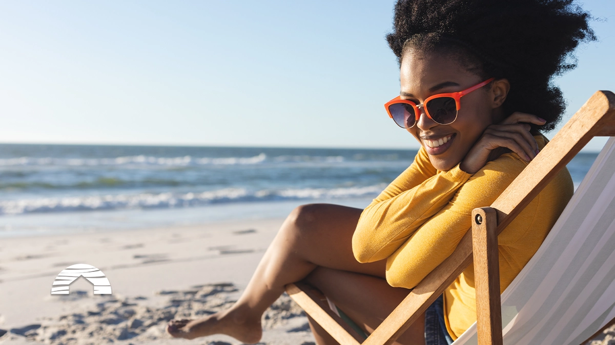 Smiling woman wearing orange sunglasses relaxing in a wooden beach chair on a sunny beach by the ocean.