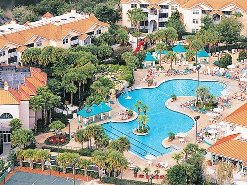 Aerial view of a large pool at Sheraton Vistana with palm trees and lounge chairs, surrounded by buildings and greenery.