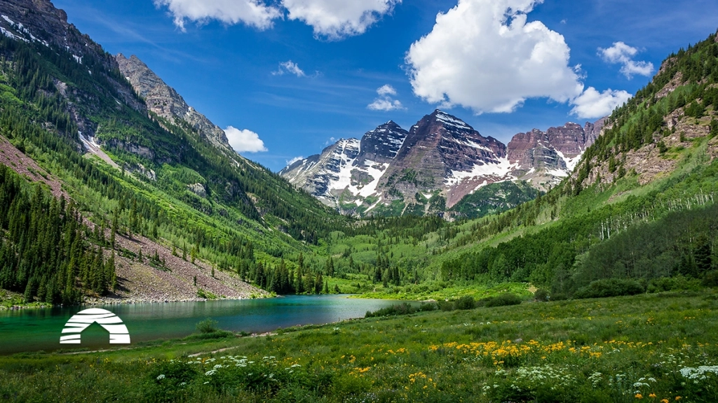 A vibrant landscape photograph of the snow-patched Maroon Bells twin peaks over the calm, deep-green Maroon Lake in Aspen, Colorado. The surrounding slopes are densely covered with pine and aspen forests, leading to a lush, foreground meadow filled with a colorful array of yellow and white wildflowers. A bright blue sky with fluffy white clouds completes the scenic Rocky Mountain view.