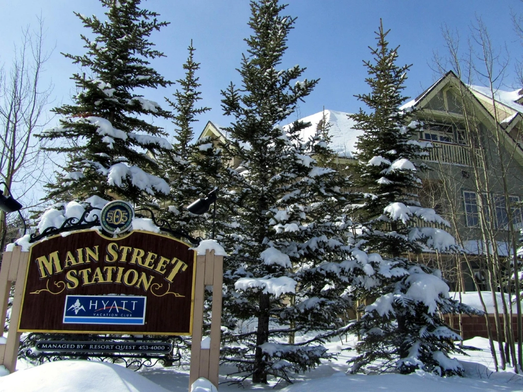 Snow-covered entrance sign for Main Street Station managed by Hyatt Vacation Club, surrounded by mature evergreen trees.