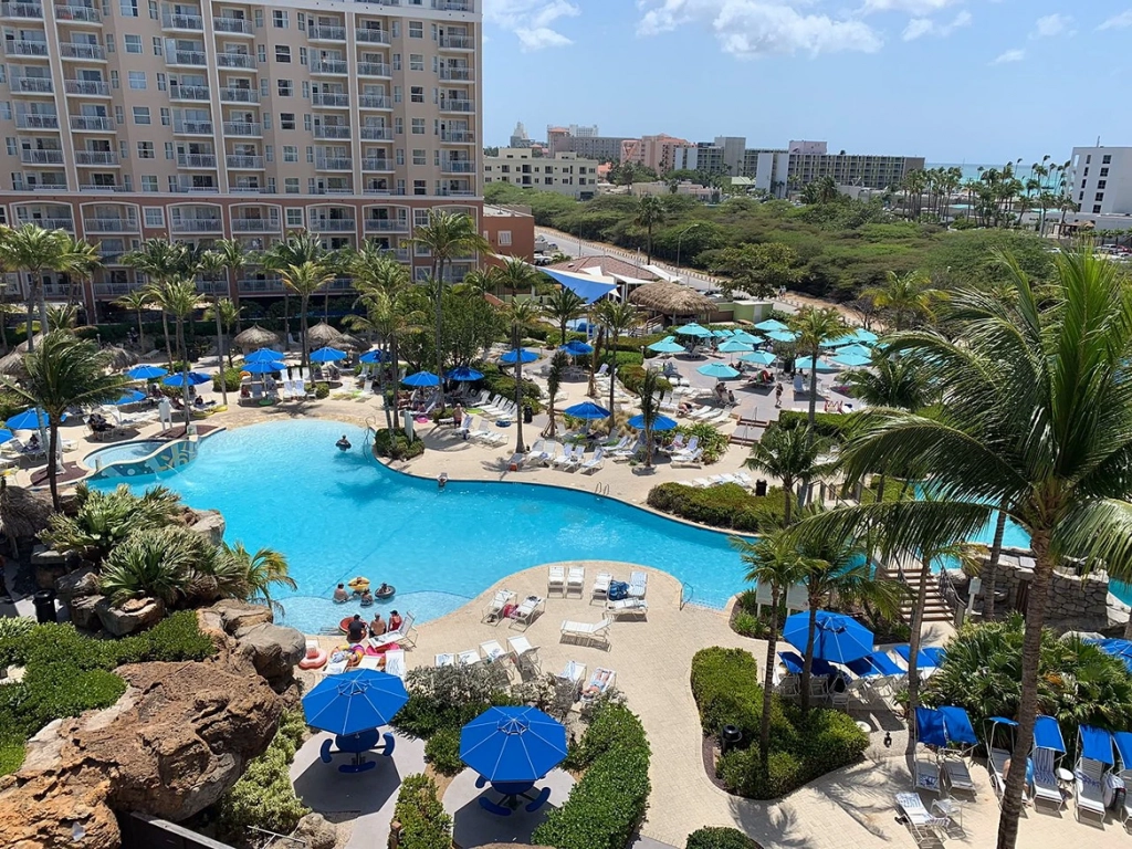 Marriott Timeshare Costs Image: A large hotel pool area at Marriott Aruba Surf Club with palm trees, blue umbrellas, and lounge chairs under a sunny sky.