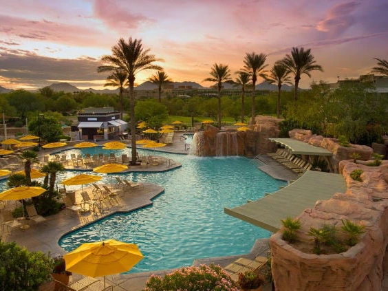 Marriott Timeshare Costs Image: A resort pool at Marriott Canyon Villas with yellow umbrellas and palm trees, surrounded by rocks under a colorful sunset.
