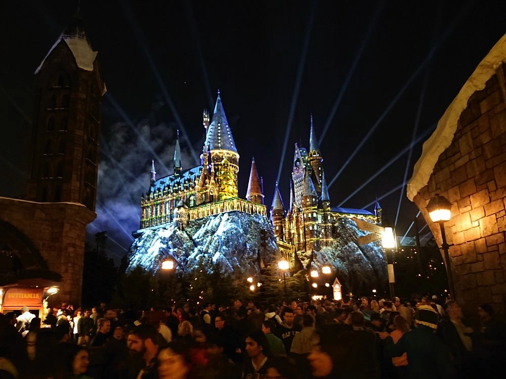 Illuminated castle on a rocky cliff at night with a light show and a large crowd of visitors gathered below.