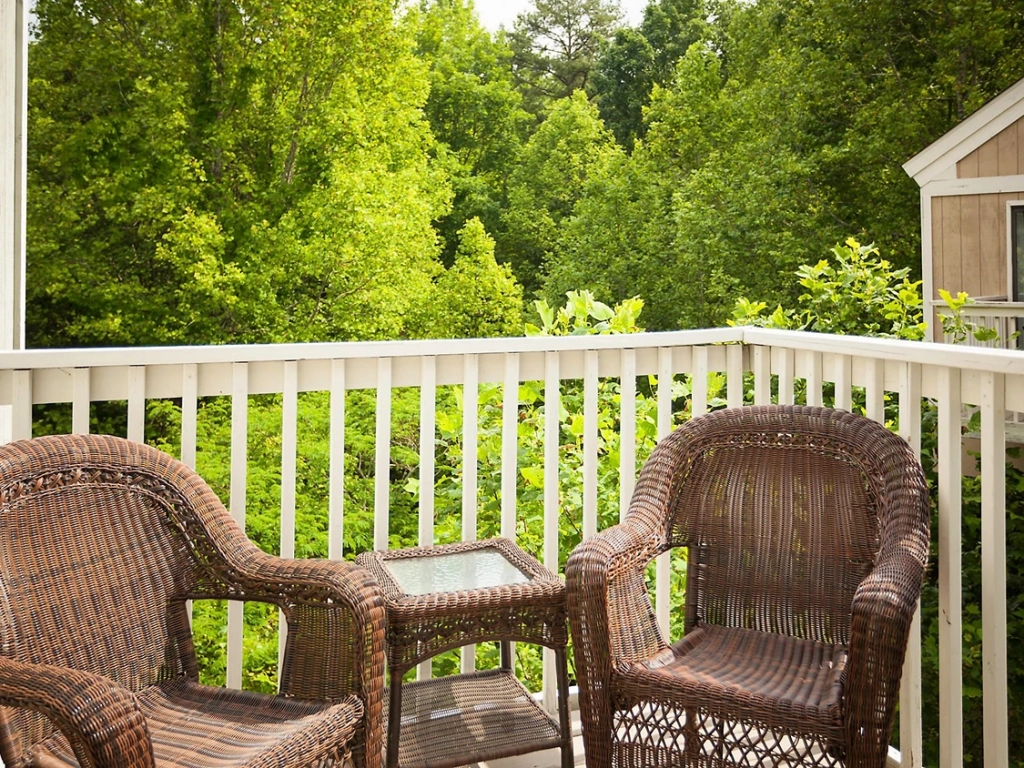 Two wicker chairs and a small glass table sit on a private balcony overlooking lush green forest trees.