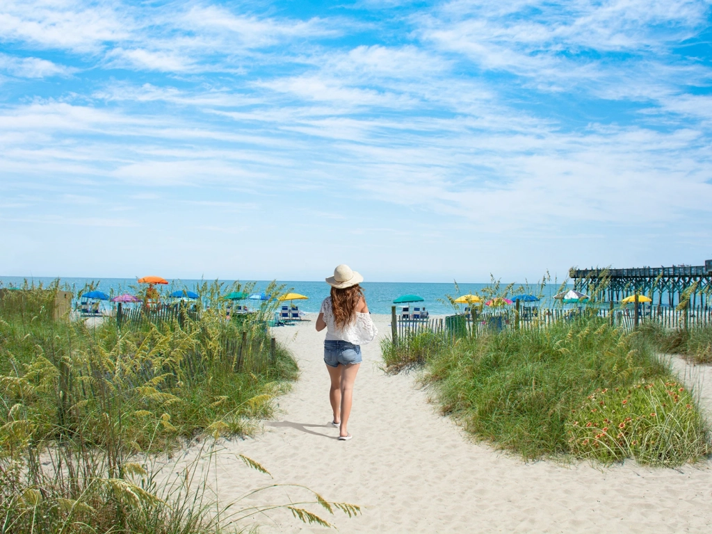 Woman in a sun hat walking down a sandy path toward the ocean and colorful beach umbrellas.