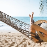 Person relaxing in a hammock on a sandy beach looking at the ocean during a summer vacation.