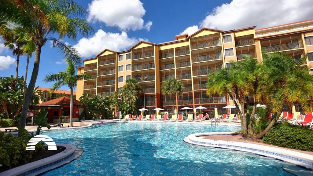 Large outdoor swimming pool with red lounge chairs and palm trees in front of a yellow resort building at Westgate Lakes.