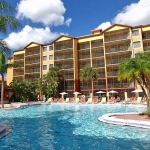 Large outdoor swimming pool with red lounge chairs and palm trees in front of a yellow resort building at Westgate Lakes.