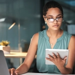 Woman researching a timeshare deedback on her tablet and laptop in a dark office.