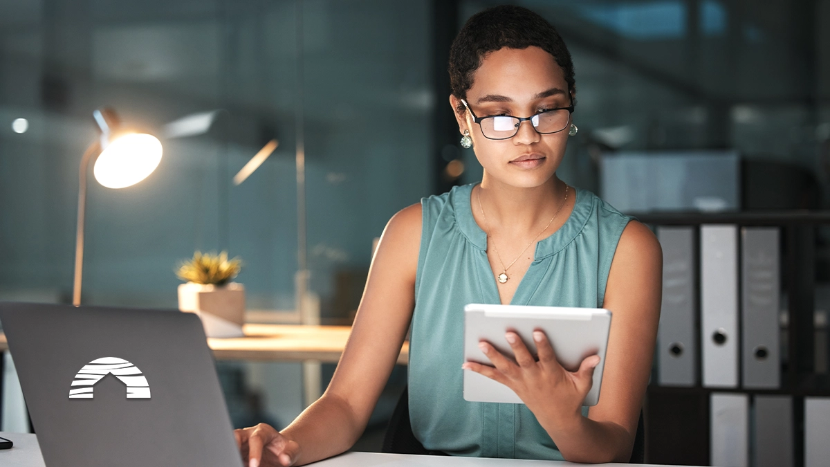 Woman researching a timeshare deedback on her tablet and laptop in a dark office.