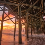 Waves washing ashore under a wooden pier at sunset with glowing orange and purple skies.