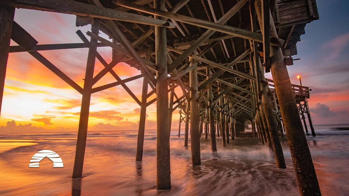 Waves washing ashore under a wooden pier at sunset with glowing orange and purple skies.