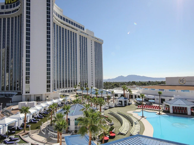 High-angle view of a large luxury resort pool deck lined with blue-roofed cabanas and lounge chairs next to a white skyscraper tower.