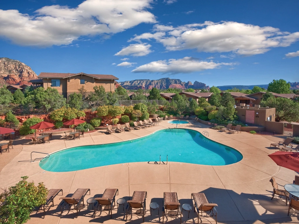 Guests relax by a large pool with red umbrellas at this Timeshare in Sedona, with red rock mountains in the background.