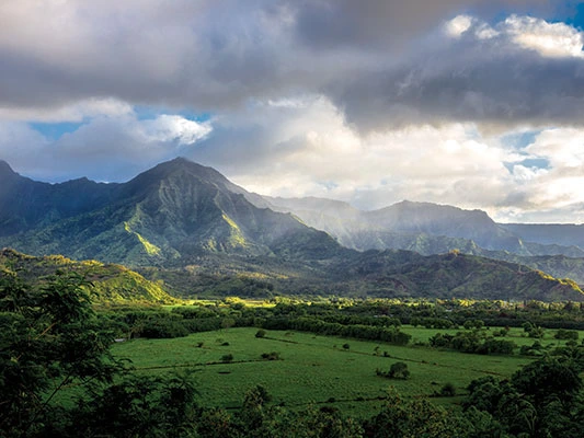 Sunlit green valley surrounded by dramatic, misty mountain peaks under a cloudy sky.