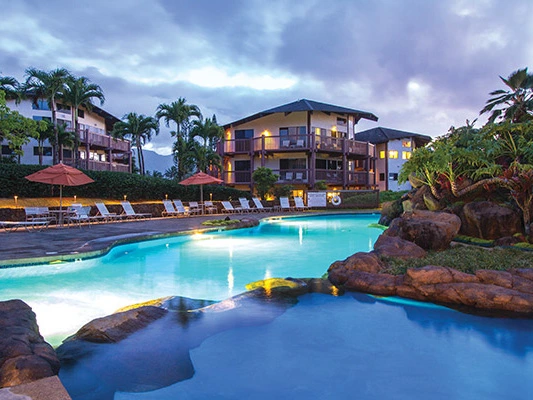 Illuminated outdoor swimming pool and lounge chairs at KaEoKai resort under a cloudy evening sky.