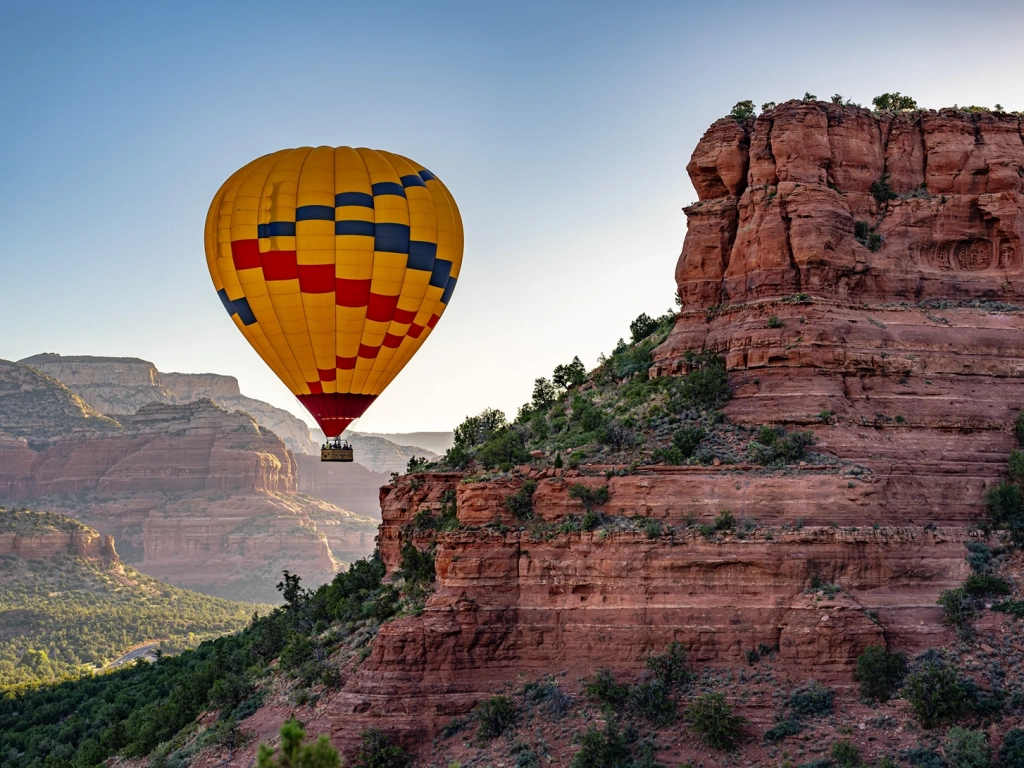 A colorful yellow, red, and blue hot air balloon floats closely past a towering, layered red rock cliff face.