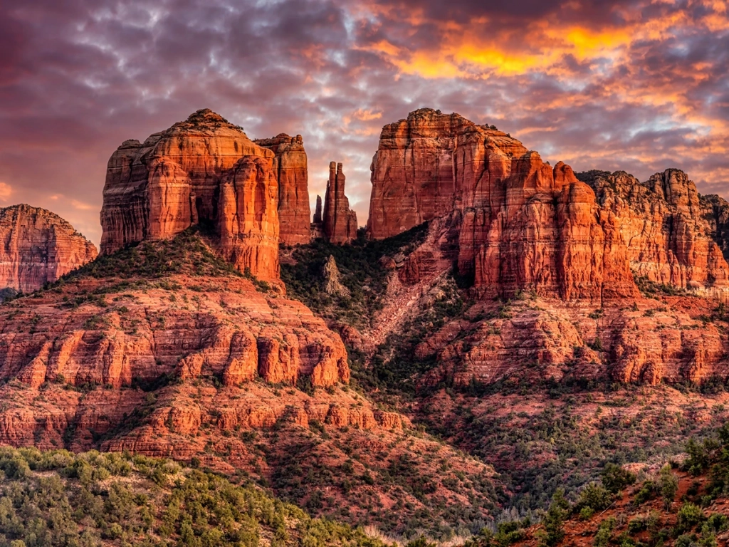 Iconic red rock mountain formations in Sedona are warmly illuminated under a vibrant sky with orange and purple clouds.