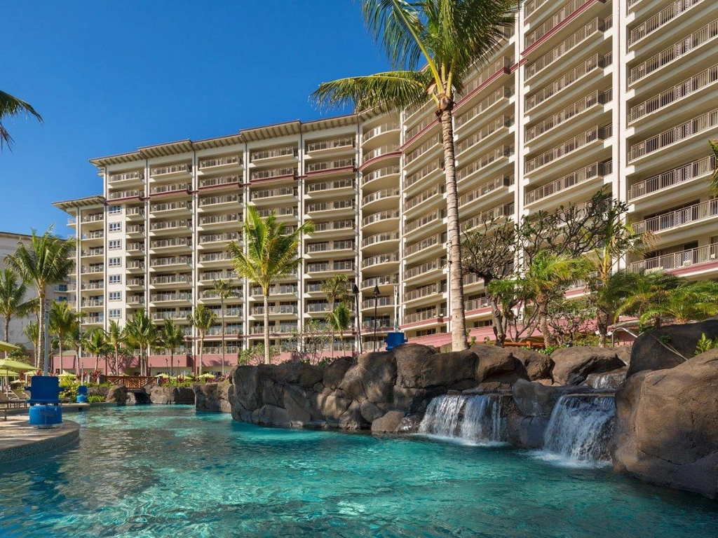A multi-story resort building with balconies overlooks a tropical lagoon pool with rock waterfalls and palm trees in Maui.