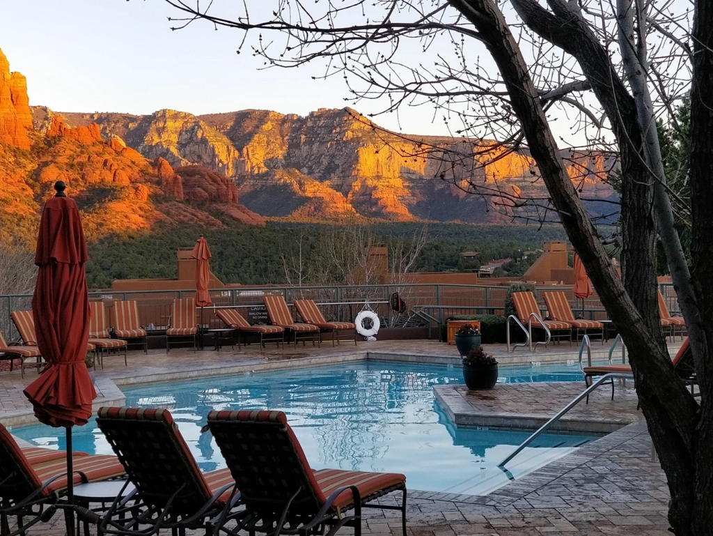 A serene outdoor pool reflects the sunlit red rock mountains at dusk, surrounded by lounge chairs and closed umbrellas.