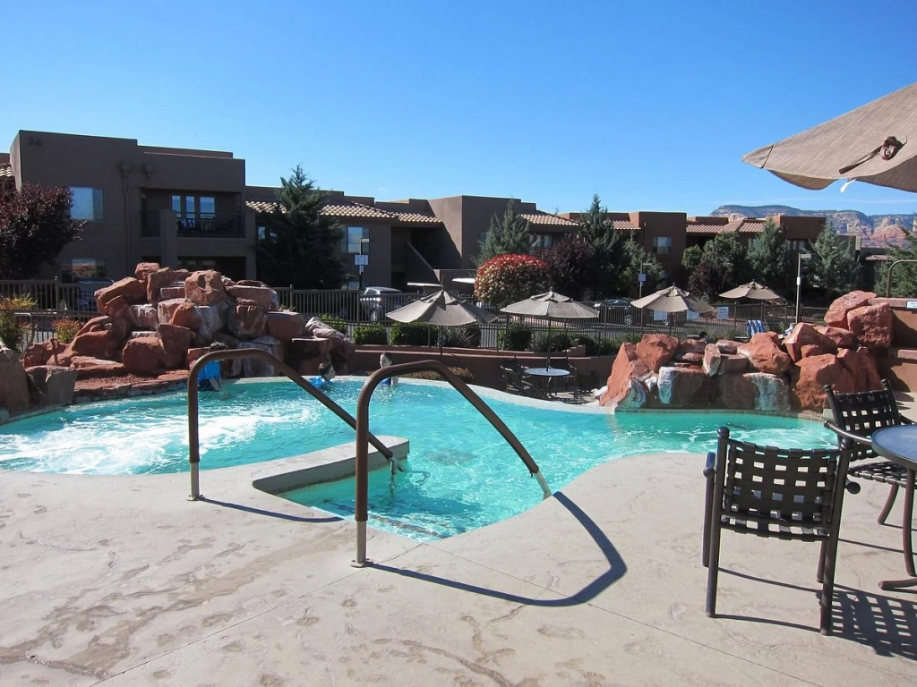 Guests relax by a multi-tiered outdoor pool and hot tub area featuring artificial red rock waterfalls.