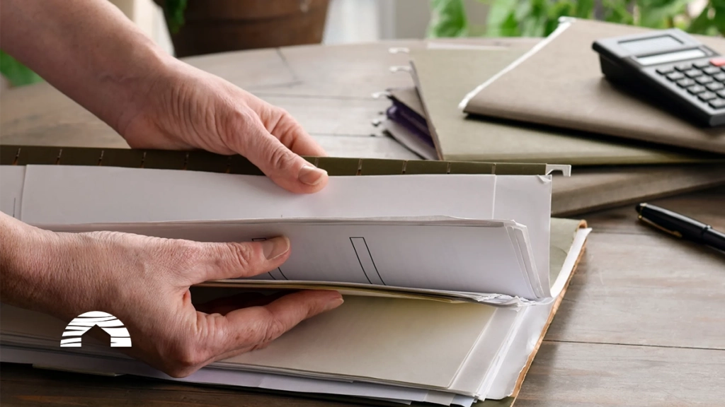 Hands opening a binder filled with documents on a wooden desk beside a calculator and pen.
