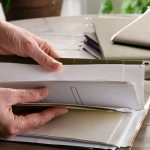 Hands opening a binder filled with documents on a wooden desk beside a calculator and pen.