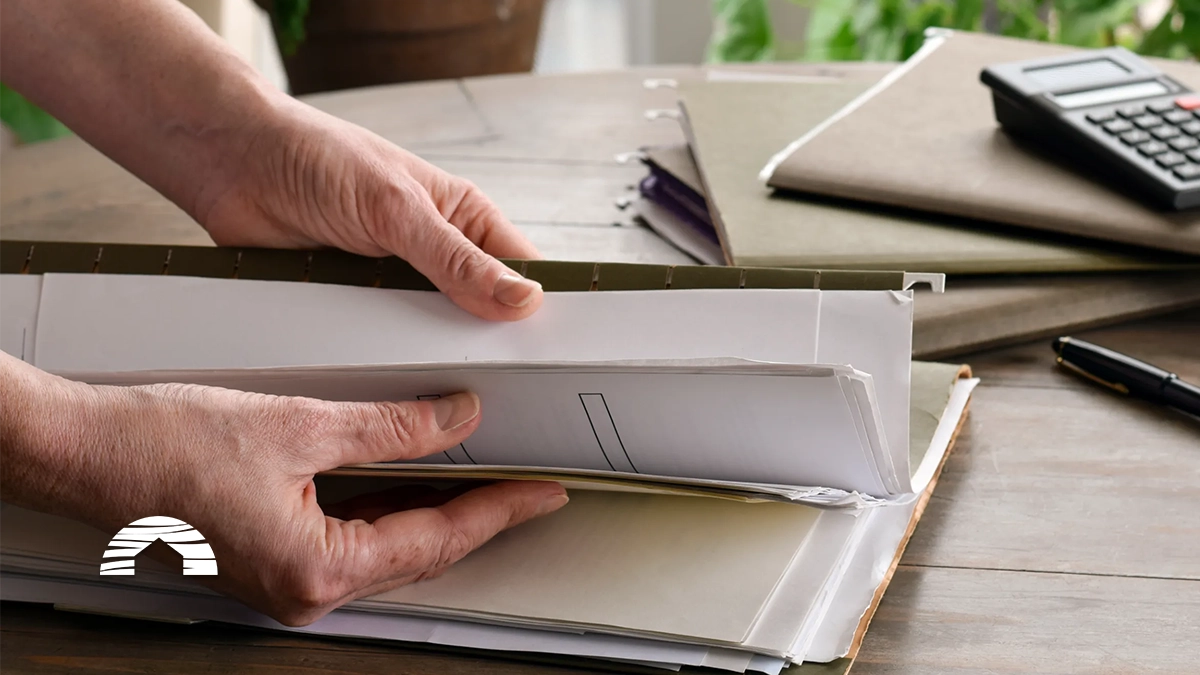 Hands opening a binder filled with documents on a wooden desk beside a calculator and pen.