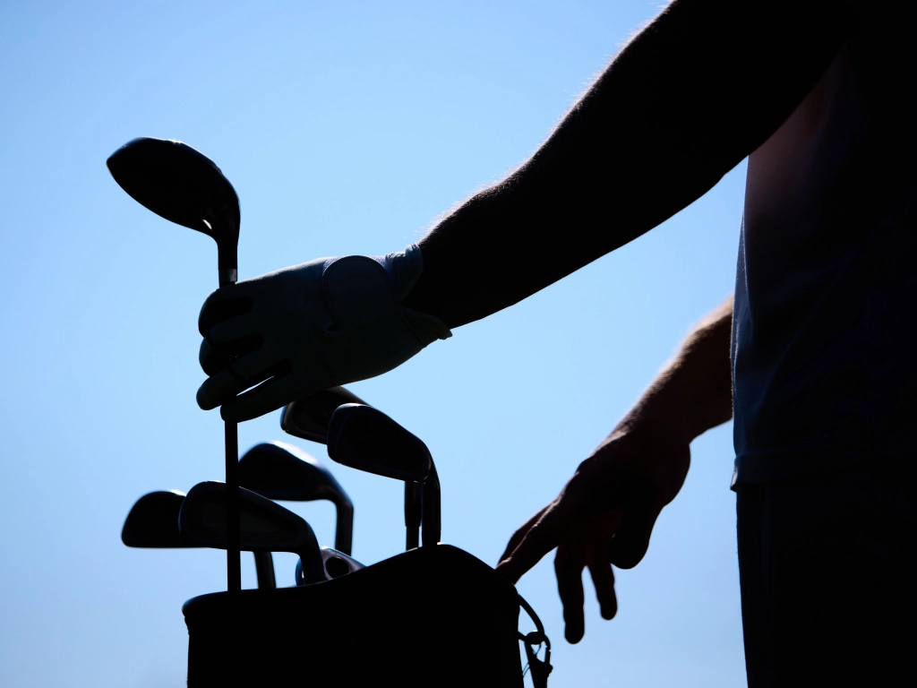 Silhouette of a golfer reaching for a club in a golf bag against a bright blue sky.