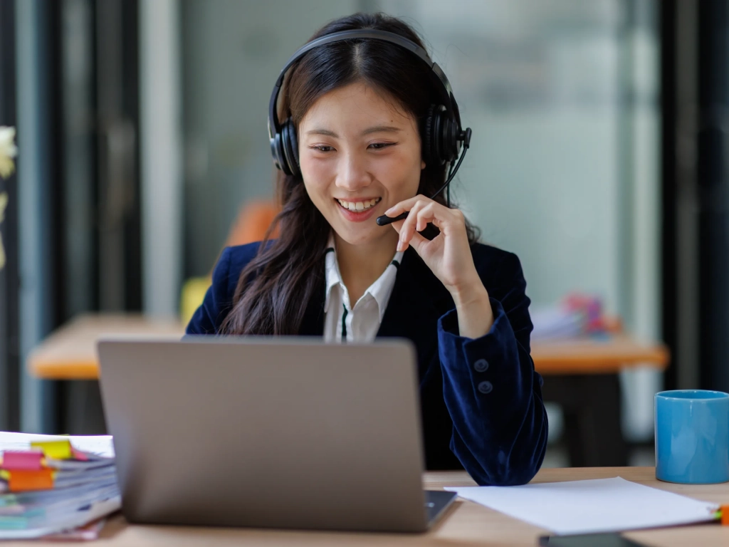 Smiling woman in a blazer wearing a headset speaks into a microphone while working on a laptop at a desk.