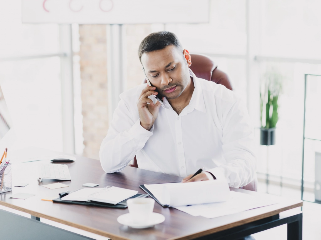 A man sitting at an office desk talking on a cell phone while writing on a notepad.