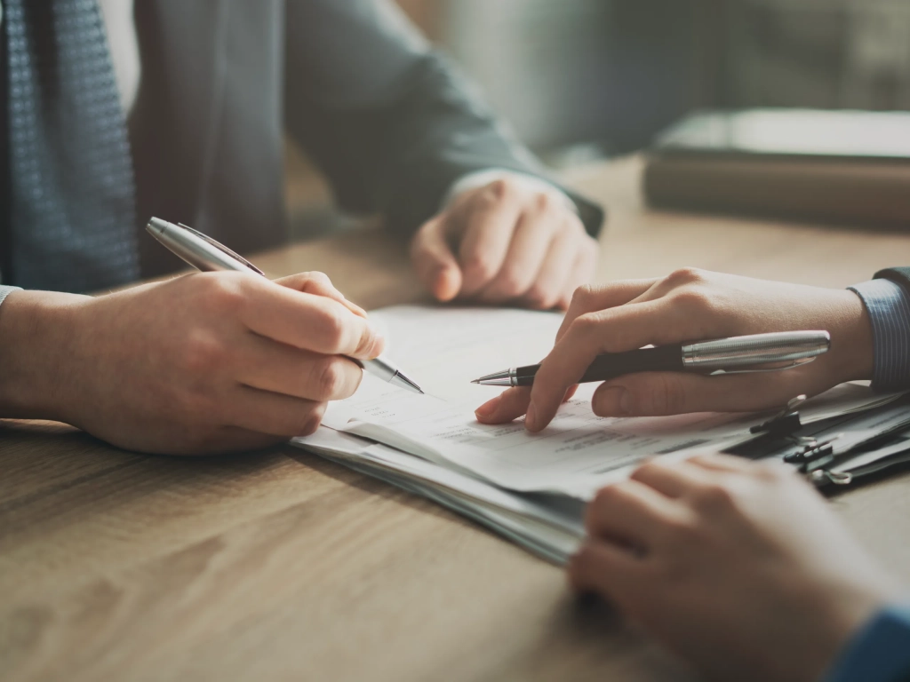 Two people signing an estoppel certificate at a wooden desk, each reviewing the document with a pen in hand.