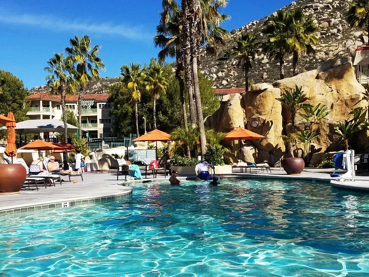 Sunny resort pool with lounge chairs and orange umbrellas at a San Diego timeshare, framed by rocky landscaping.
