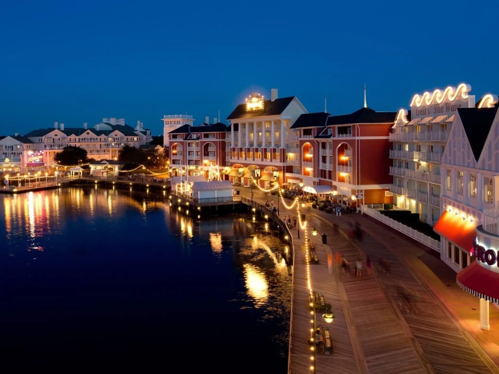 Illuminated boardwalk and waterfront buildings reflected in calm water at dusk near Disney's BoardWalk Resort.