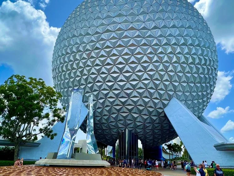 Spaceship Earth geodesic sphere at EPCOT rises above visitors and a reflective crystal monument on a sunny day.