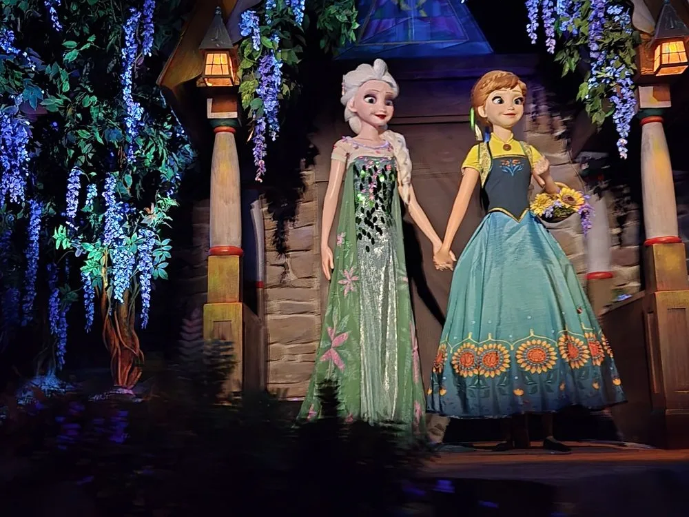 Elsa and Anna animatronics on one of the most beloved Epcot rides, holding hands beneath glowing wisteria and lanterns.