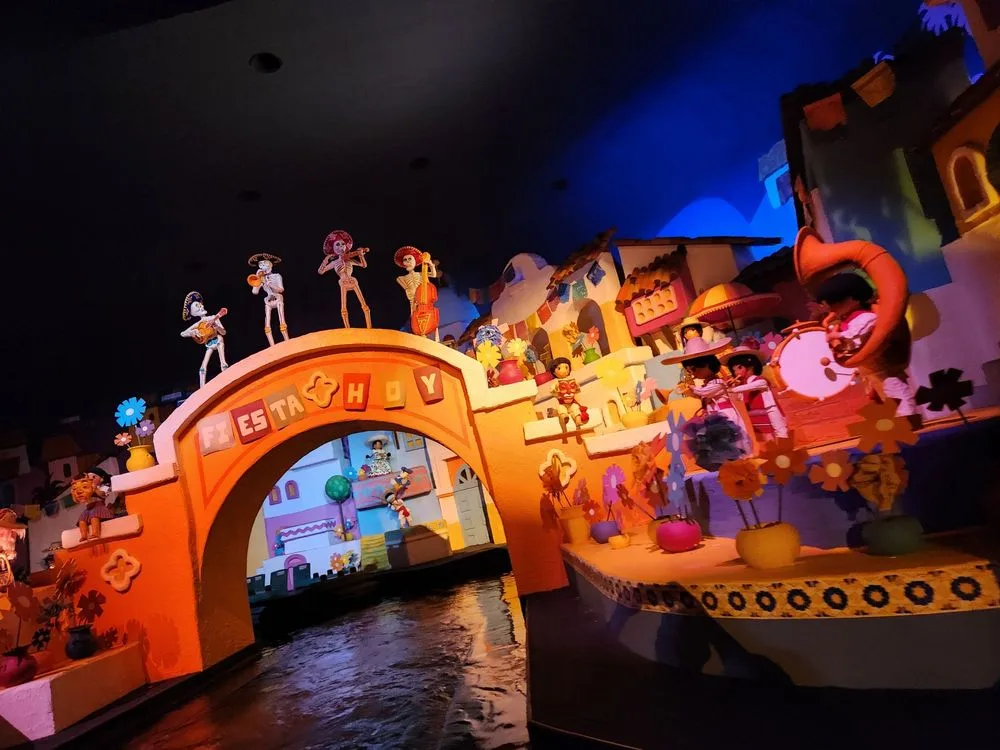 Colorful Day of the Dead skeleton musicians perform atop an arched bridge in a dark indoor boat ride scene.