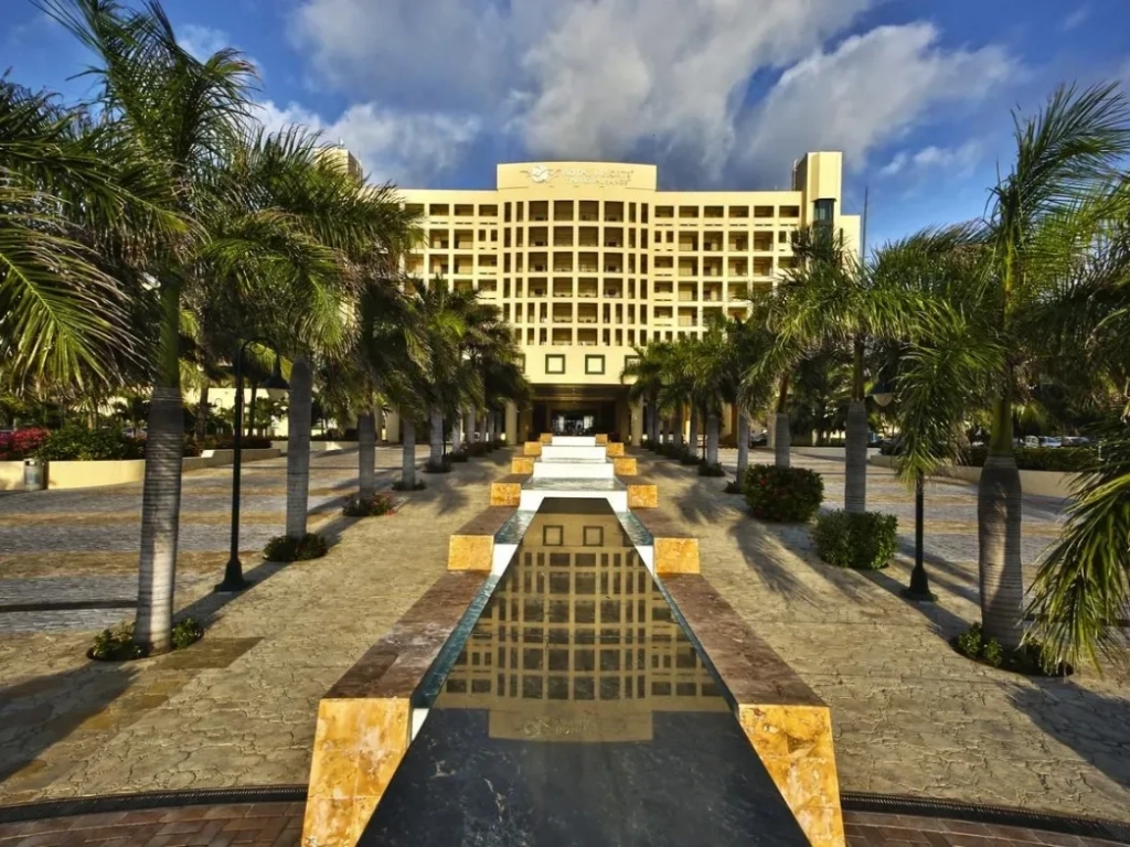 Grand resort hotel with yellow facade viewed from a symmetrical palm-lined entrance walkway with a reflecting pool.