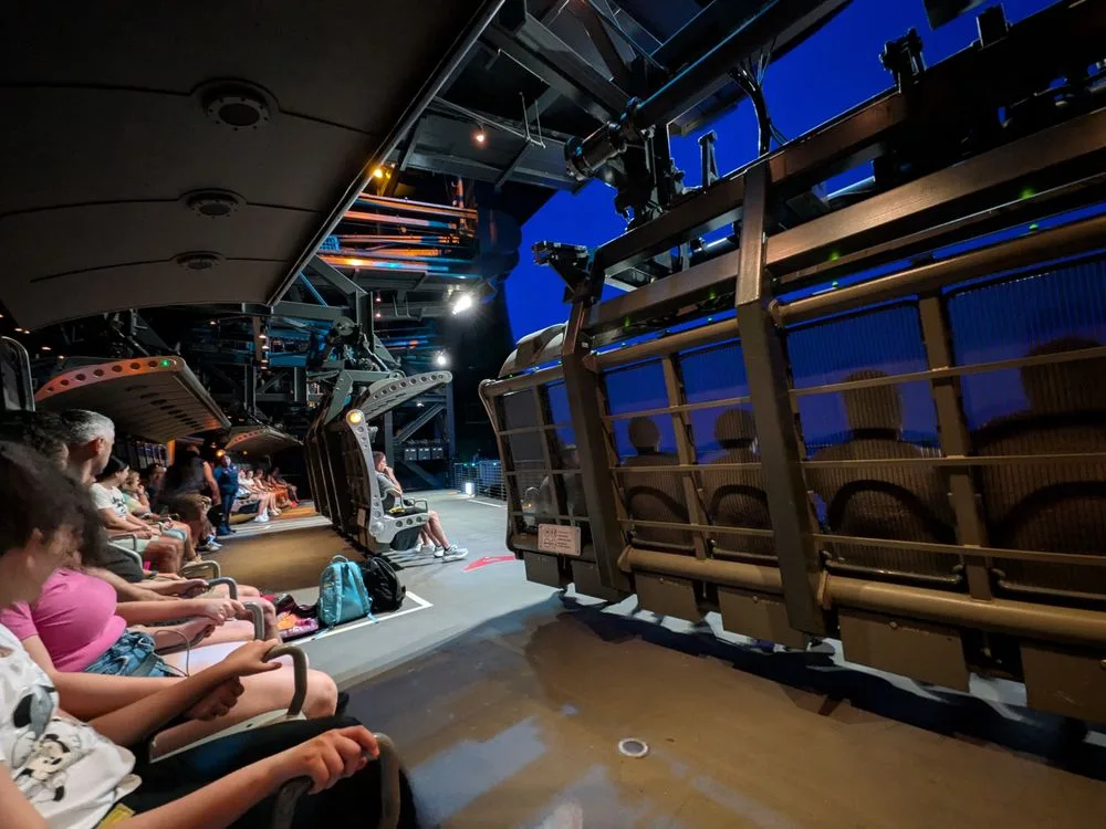 Guests seated in rows wait to board a large gondola-style ride vehicle at a darkened indoor theme park attraction.