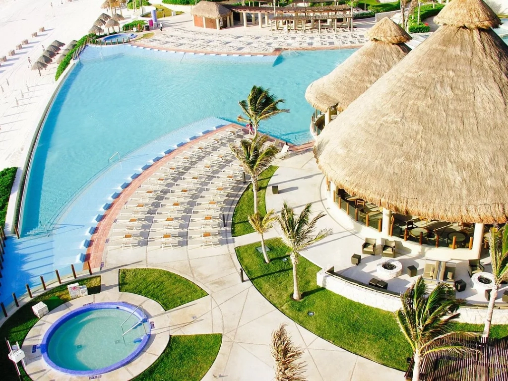Aerial view of a large curved resort pool at a timeshare in cancun, with lounge chairs, palm trees, a hot tub, and thatched-roof bar pavilion.